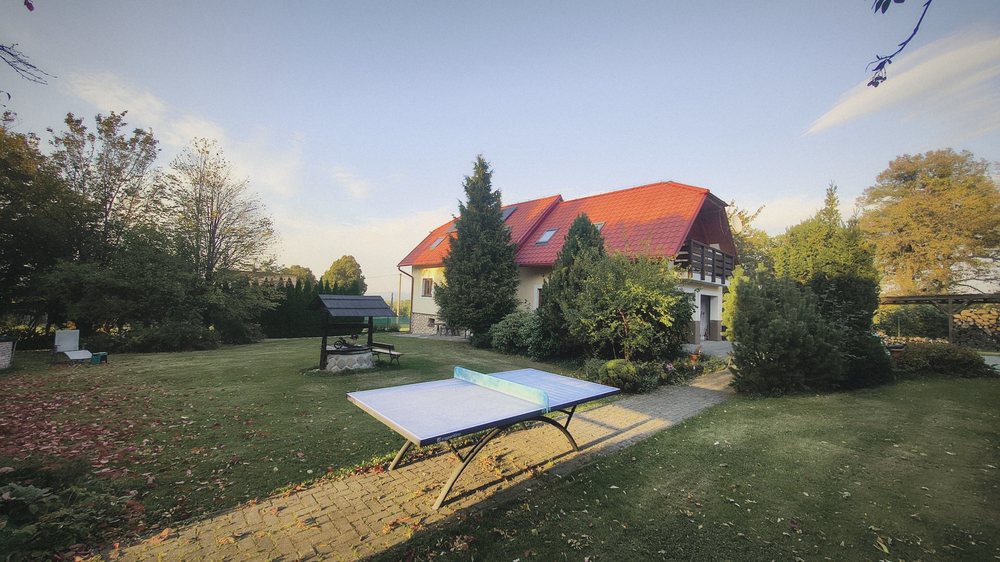 Chalupa s červenou střechou a balkonem, před ní zahrada se stolem na ping-pong a studnou